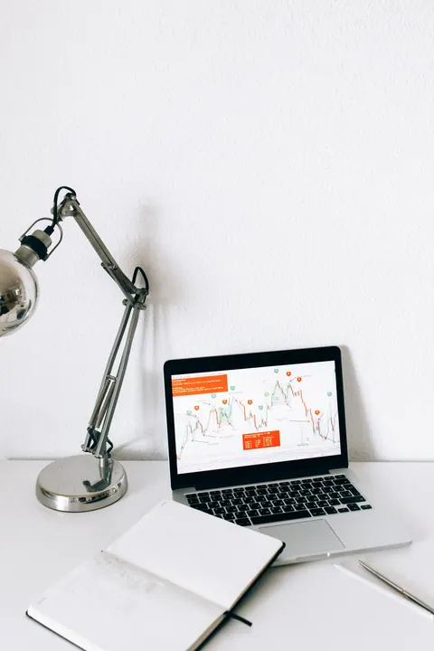 Laptop displaying charts beside a notebook and desk lamp