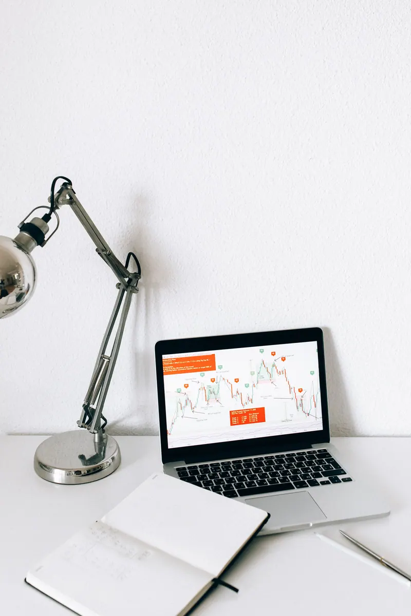 Laptop displaying charts beside a notebook and desk lamp